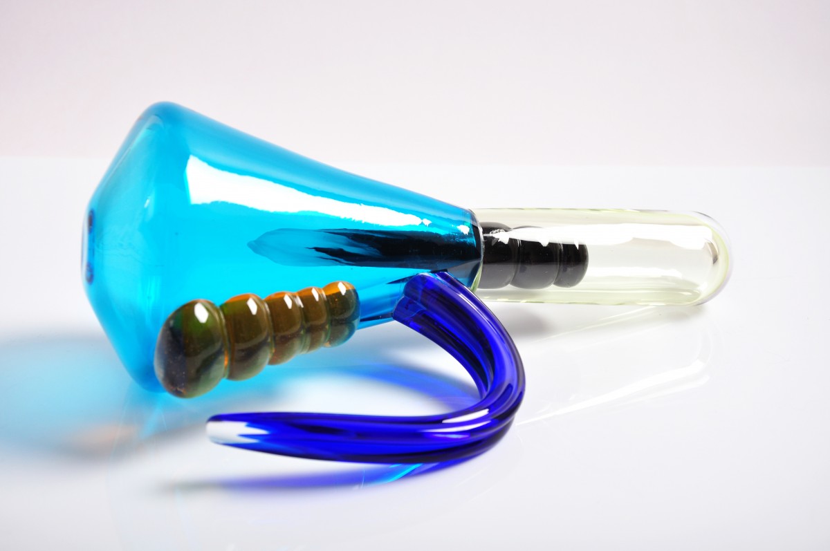 G;ass sculpte that looks like a ray gun. A bright blue tapered glass shape attaches to a pale green tube. Inside there is an opaque black section and on the base is a series of yellow bumps and a darker blue curved handle.