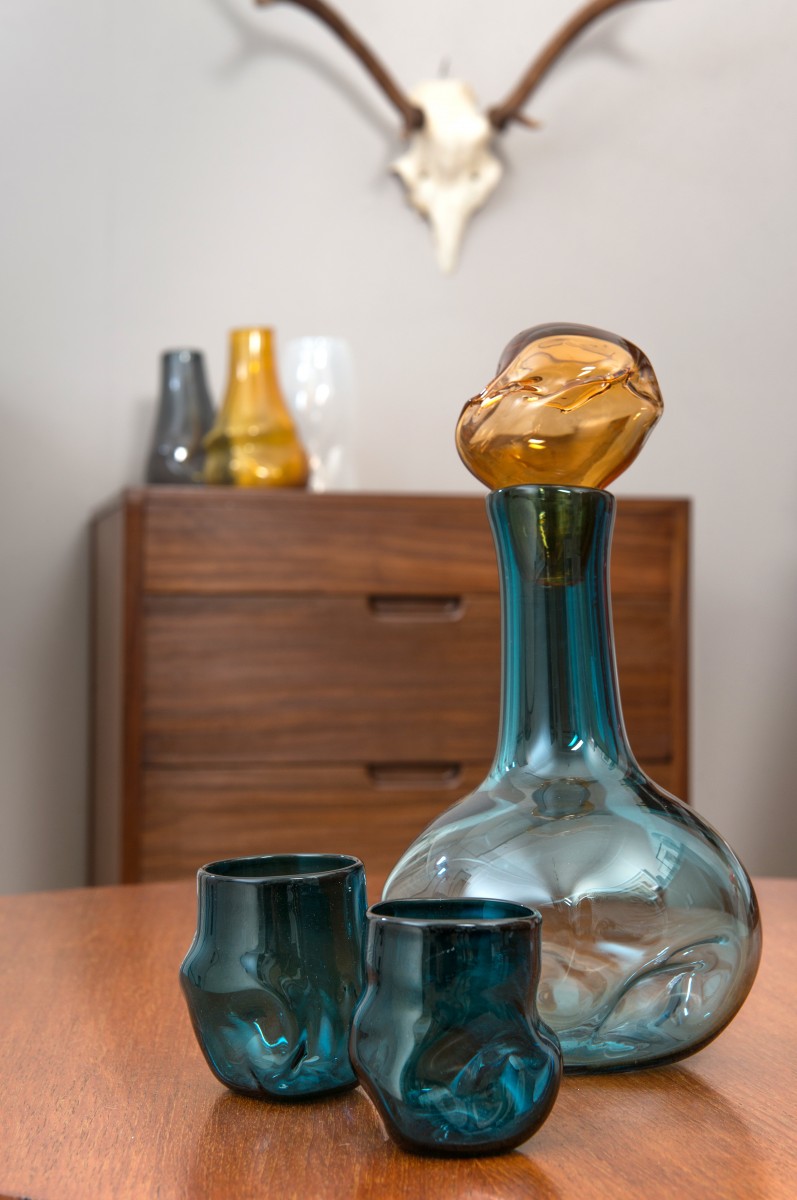 Blue glass decanter and glasses with apricot stopper in front of drawers with glass vases and stag skull