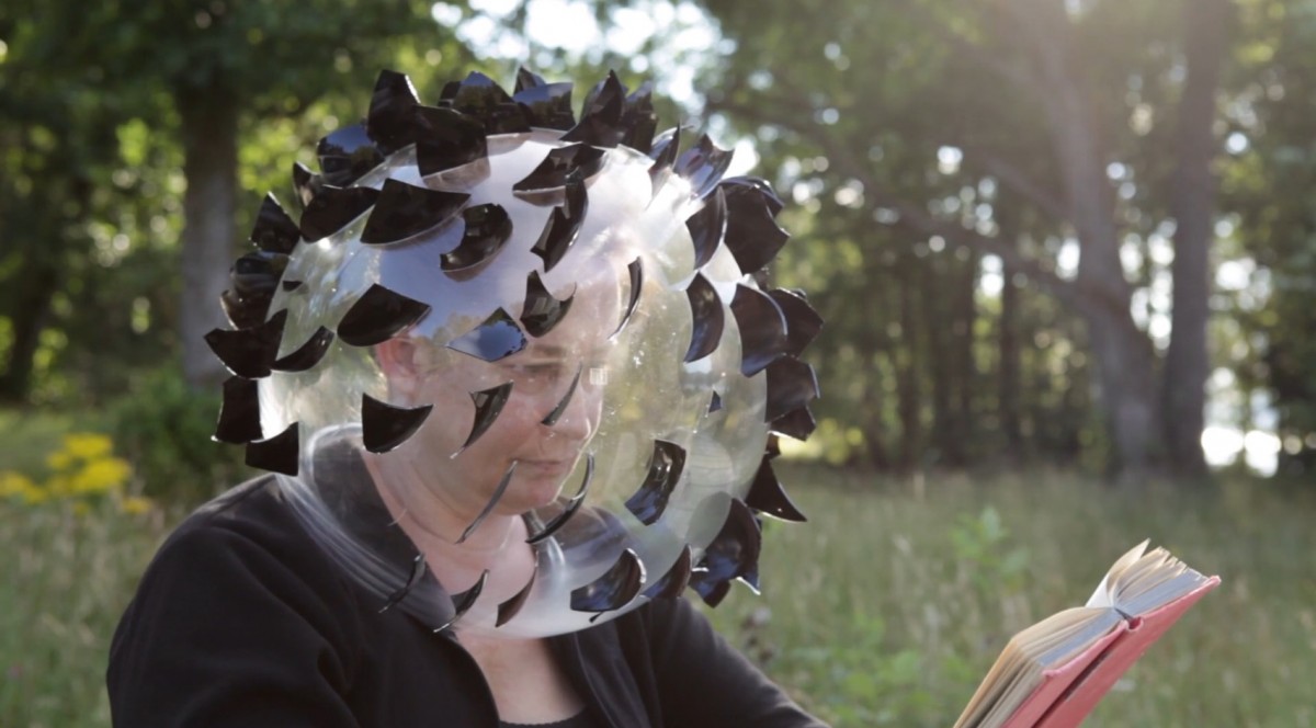 Still from a video showing the artist reading a book whilst wearing a clear glass dome over her head, covered in black glass shards.